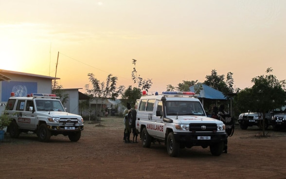 Deux véhicules tout-terrain blancs portant les inscriptions "UN" et "Ambulance" sont garés sur une place sablonneuse.